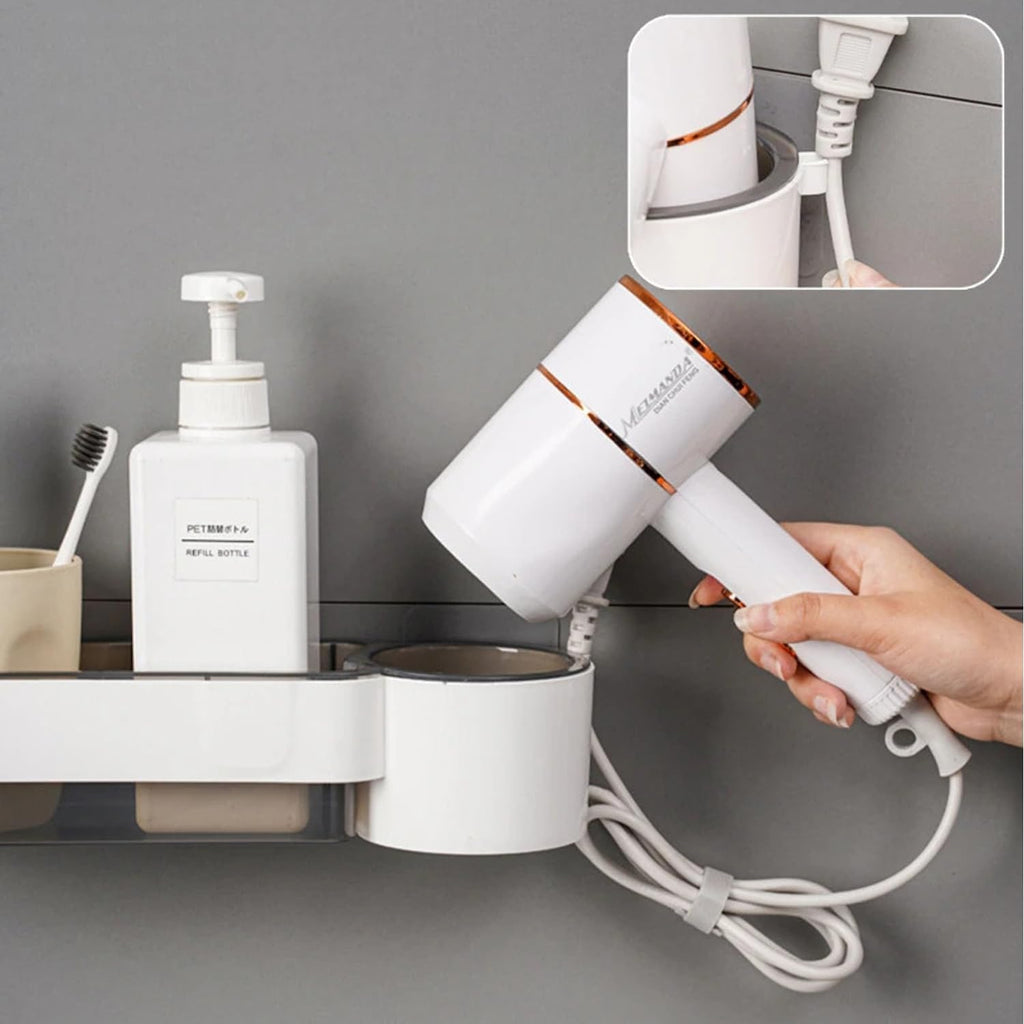 A white hair dryer with metallic copper accents is being held by a hand against a gray bathroom wall. A wall-mounted white holder is visible, along with a toothbrush holder and soap dispenser.