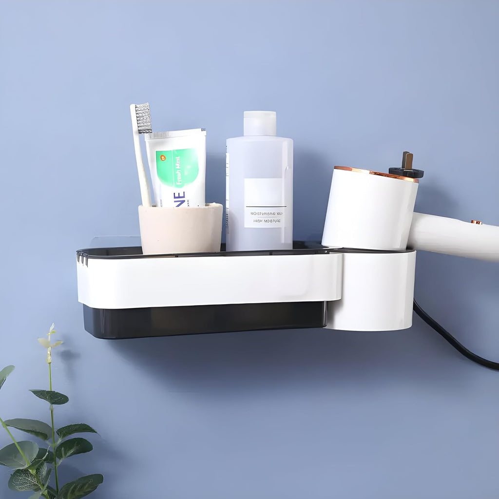 A wall-mounted bathroom organizer shelf with a cup, toothbrush, toothpaste, and a bottle, attached to a white and black wall-mounted hair dryer. The setup is mounted on a light blue wall with a small plant in the lower left corner.