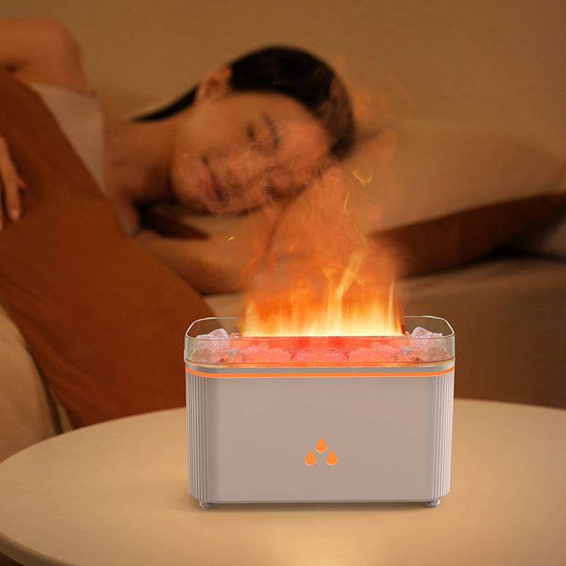 A tabletop flame diffuser with a glowing orange flame effect and pink stones inside, placed on a table in a bedroom setting with a blurred person sleeping in the background.