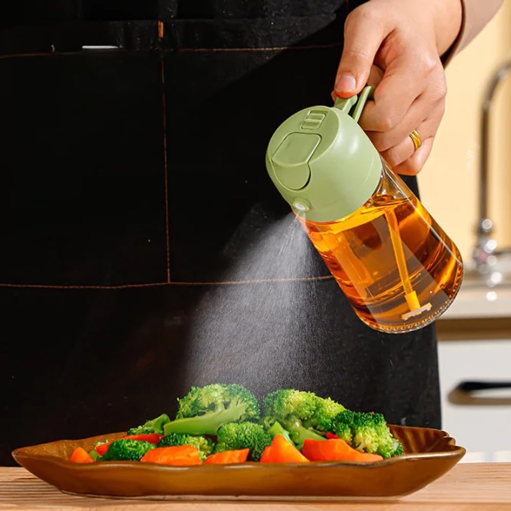 A hand holds a green and clear oil sprayer bottle above a plate of broccoli and carrots, spraying oil mist onto the vegetables. The person is wearing a black apron in a kitchen setting.