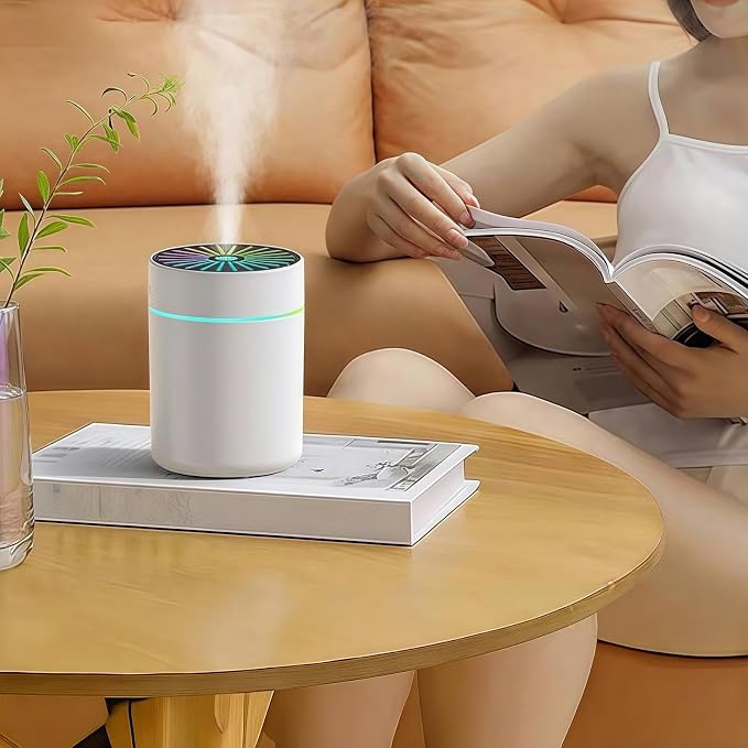 A white cylindrical humidifier with a green and blue patterned top and a glowing blue light band, emitting a mist of water. It is placed on a light wooden table next to a glass vase with a green sprig, with a woman in a white tank top sitting on an orange couch in the background reading a book.