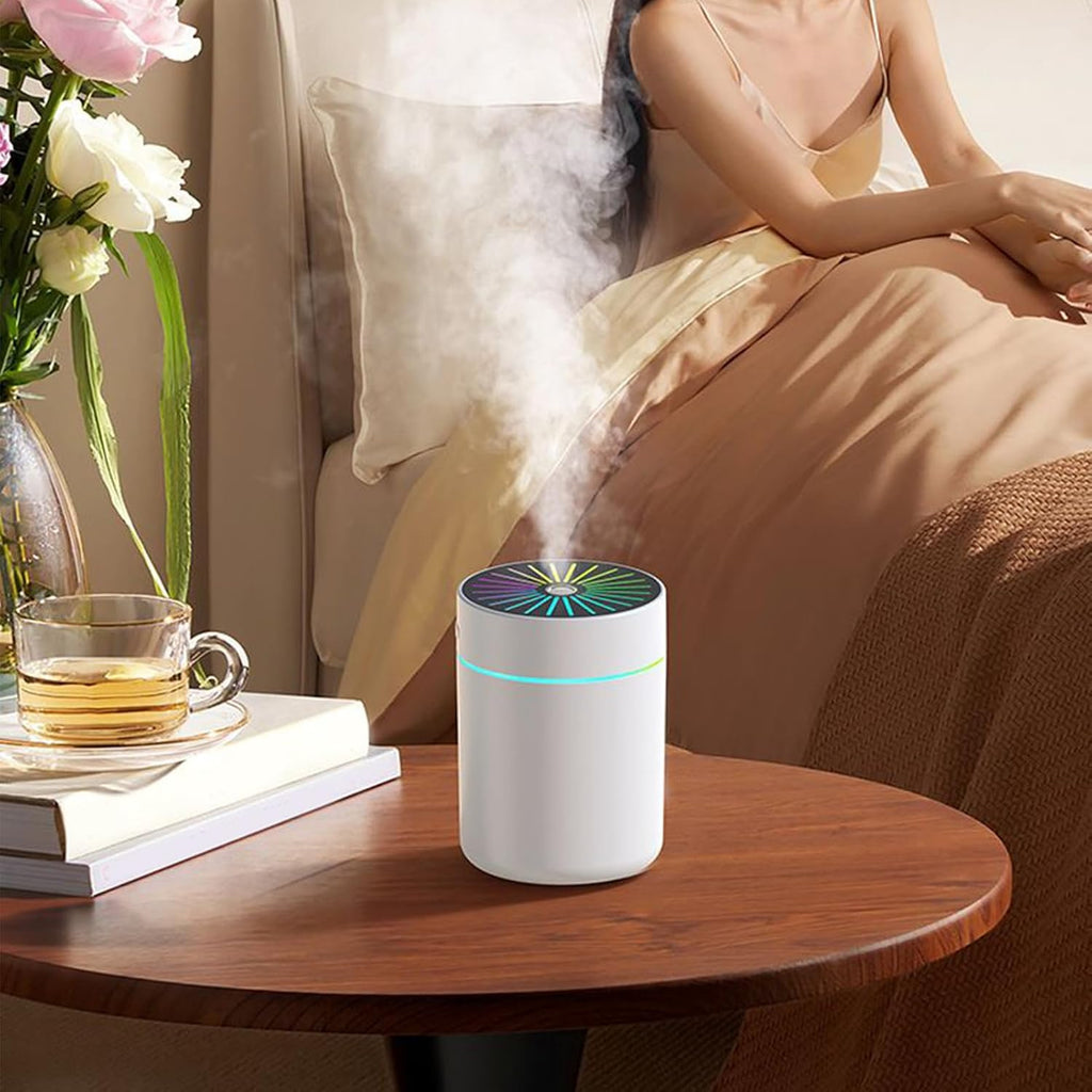 A white cylindrical humidifier emitting a plume of steam sits on a wooden bedside table next to a bed, with a woman in beige pajamas sitting in the background.