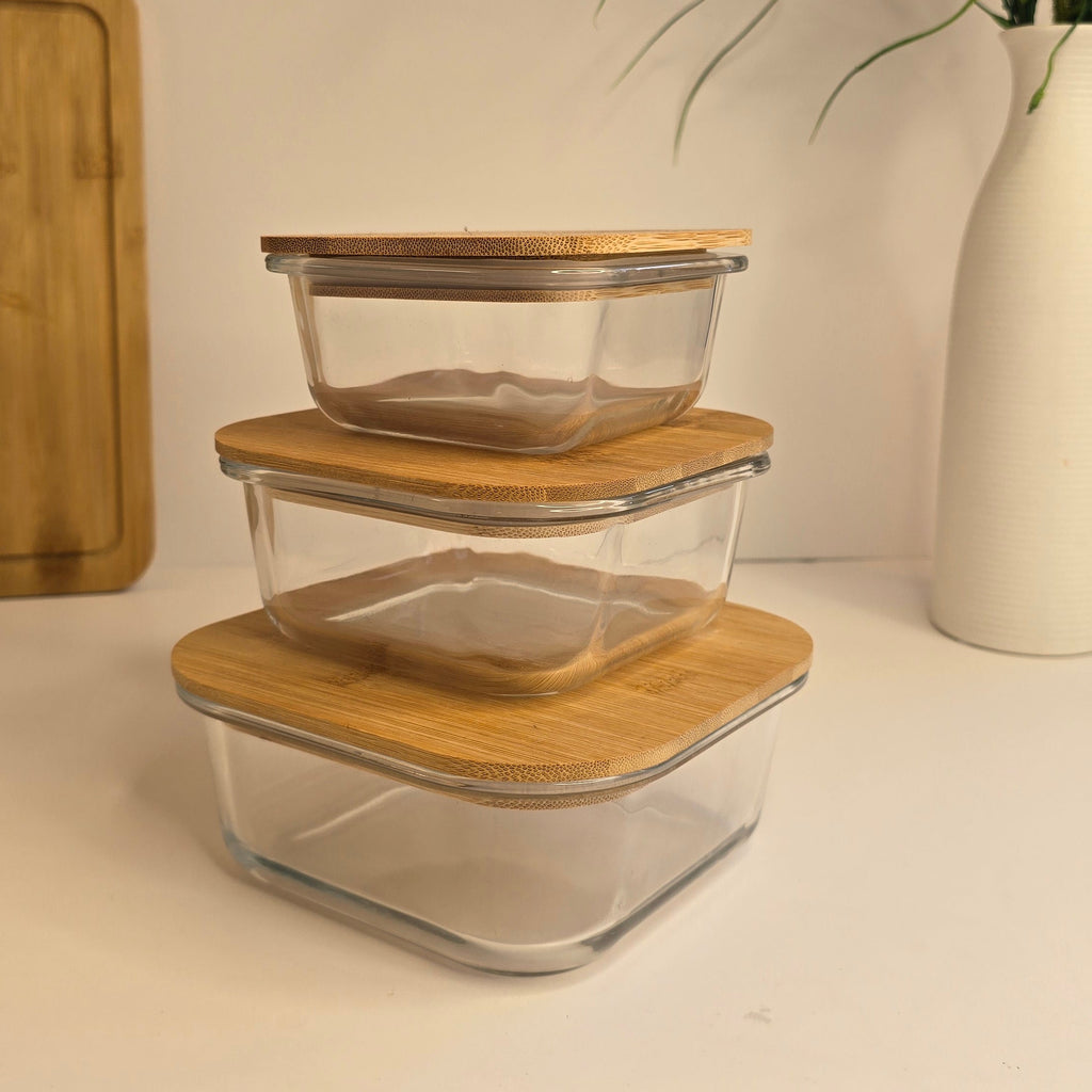 Three stacked square glass storage containers with light bamboo lids on a white surface, with a white vase of greenery and a wooden cutting board in the background.