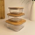 Three stacked square glass storage containers with light bamboo lids on a white surface, with a white vase of greenery and a wooden cutting board in the background.