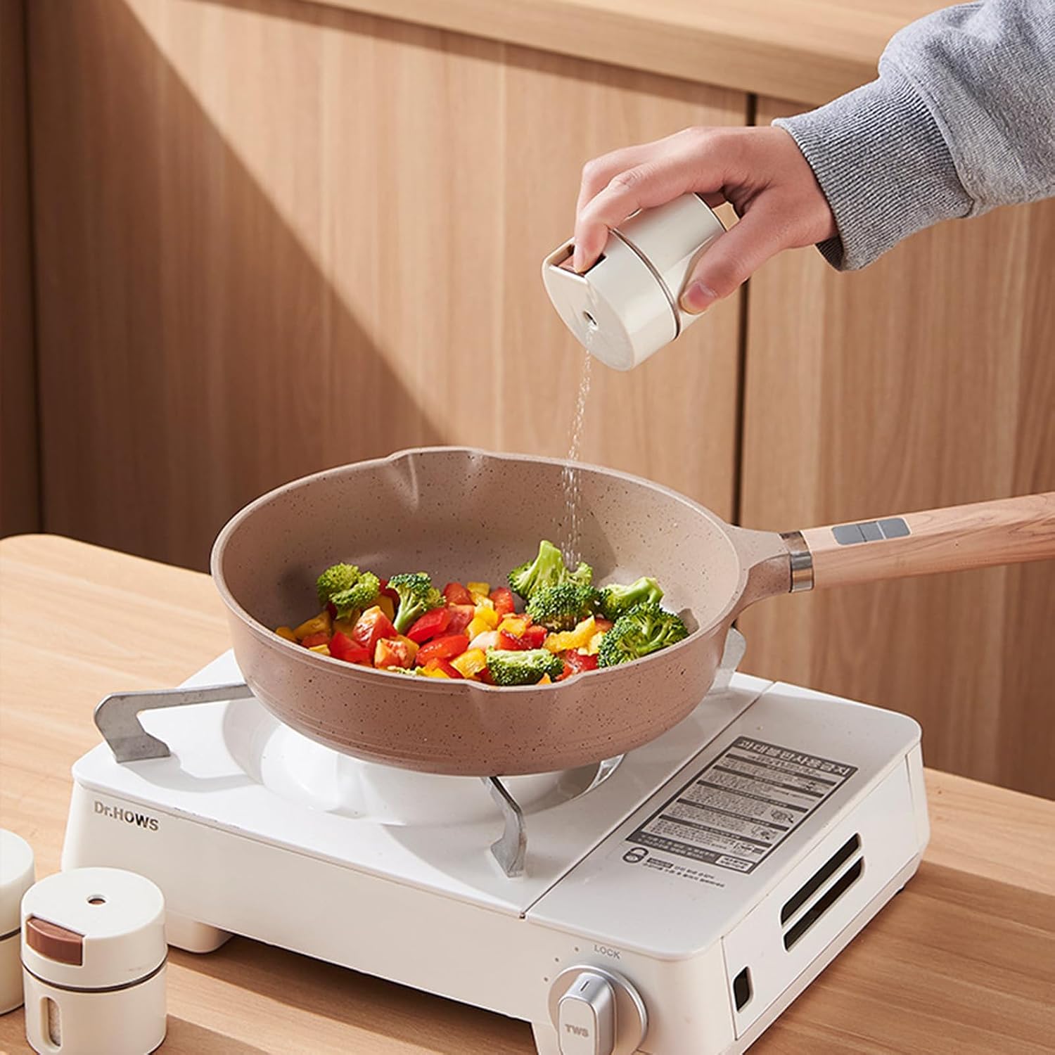 A beige non-stick frying pan with a wooden handle sits on a white portable gas stove, with a hand pouring salt from a matching white shaker container. The pan contains cooked broccoli, red peppers, and yellow peppers.