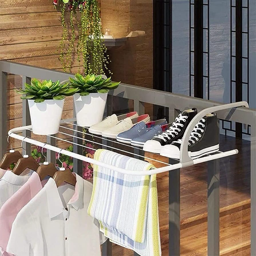 A white metal clothes and shoes rack mounted on a balcony railing, shown with folded white shirts, a striped towel, two potted green plants, and several pairs of shoes.