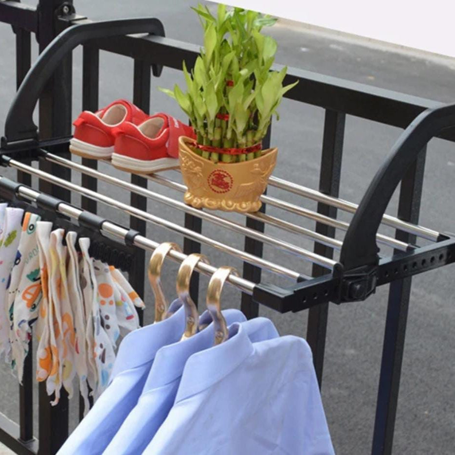 A black metal outdoor clothes rack mounted on a black metal railing, shown holding several light blue shirts on gold hangers, patterned towels, red shoes, and a potted green plant.