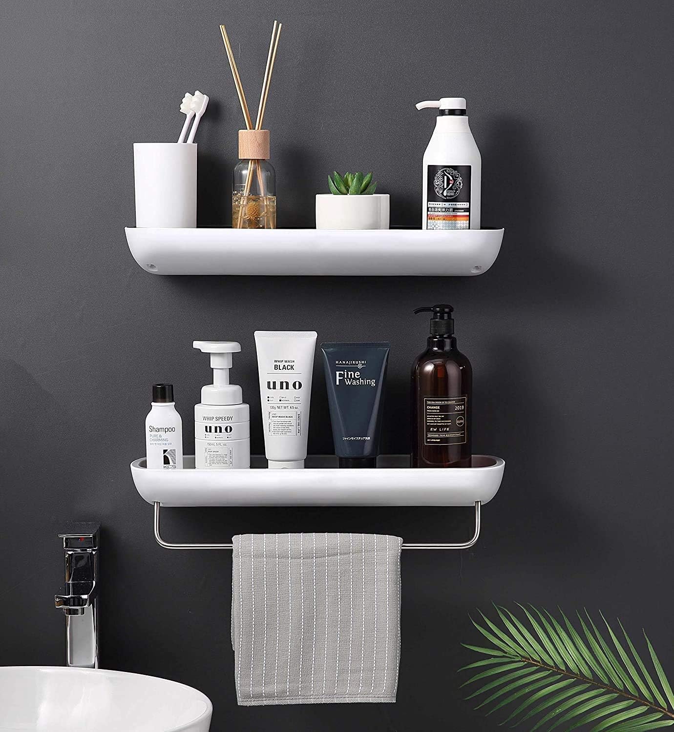 Two white wall-mounted bathroom shelves holding toiletries and a towel, installed on a dark gray wall above a sink. Visible products include a diffuser, toothbrushes in a cup, a small plant, a pump bottle, and several skincare bottles and tubes.