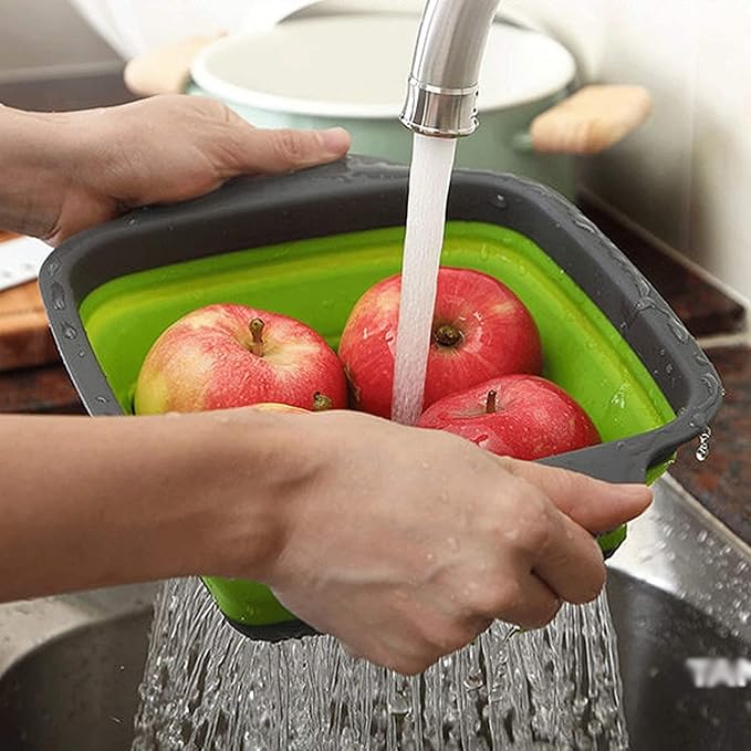A green and gray collapsible sink basket being held by hands under running water with red apples inside, set in a kitchen sink environment.