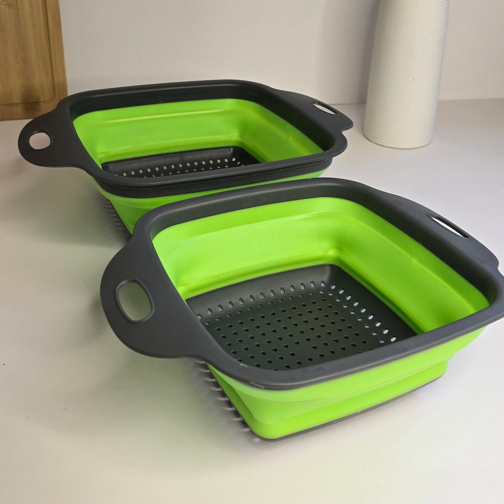 Two collapsible rectangular colanders with bright green interiors and dark gray perforated exteriors, shown on a white tabletop with a vase and dried stems in the background.