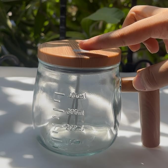 A clear glass measuring mug with a light brown wooden lid and handle, shown outdoors on a sunlit white surface with a person's hand touching the lid.