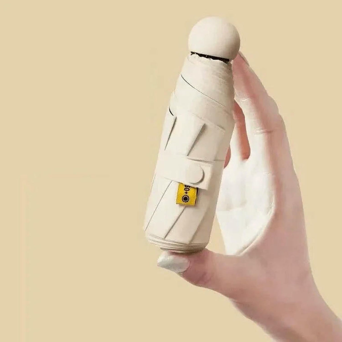 A hand holding a compact umbrella wrapped in off-white fabric with a small yellow tag, set against a solid beige background.