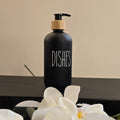 A matte black pump bottle labeled 'DISHES' with a light wood collar, placed on a dark shelf in front of white orchid flowers against a beige wall.