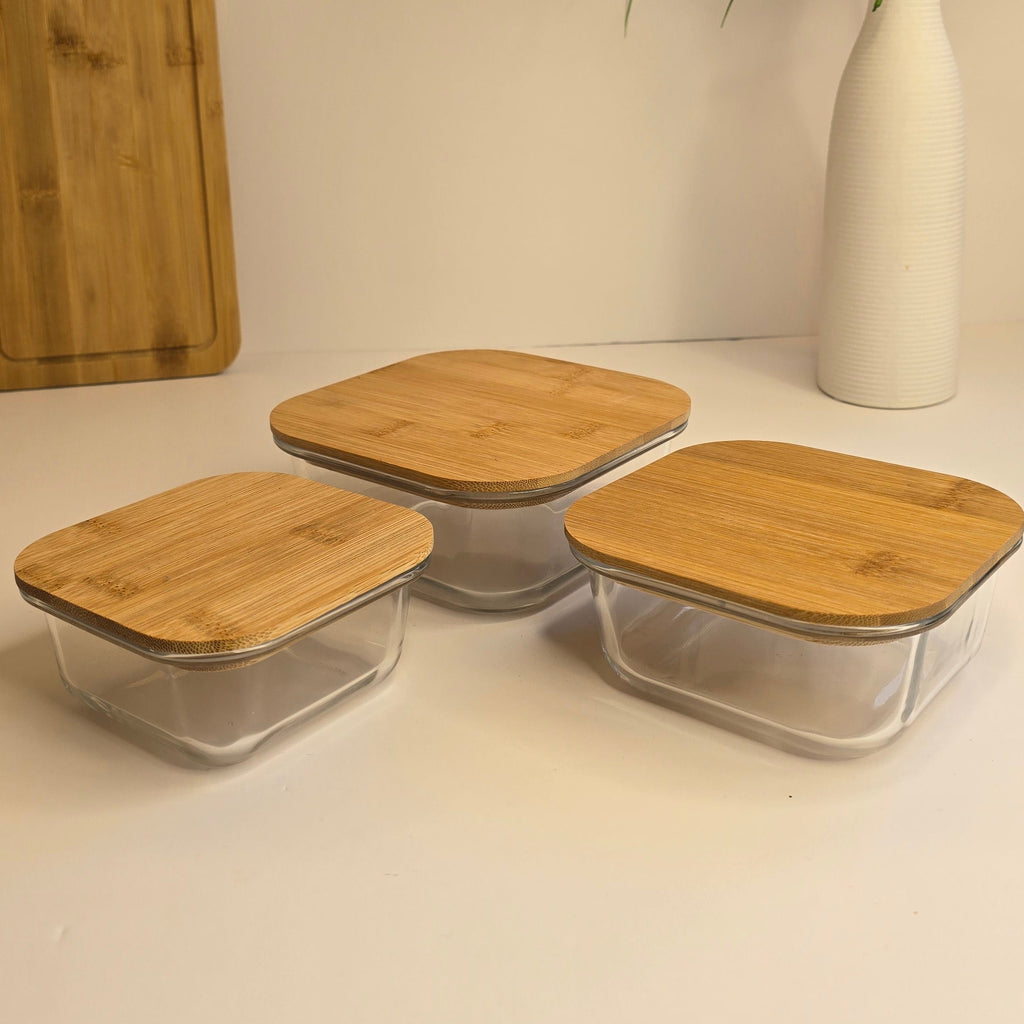 Three clear glass storage containers with square light wood lids arranged on a white surface, with a white vase of flowers and a wooden cutting board in the background.