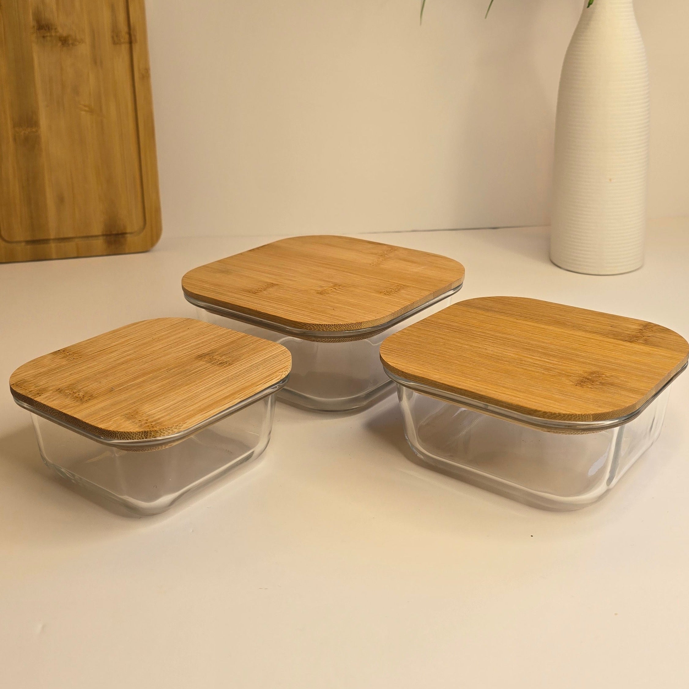 Three clear glass storage containers with square light wood lids arranged on a white surface, with a white vase of flowers and a wooden cutting board in the background.