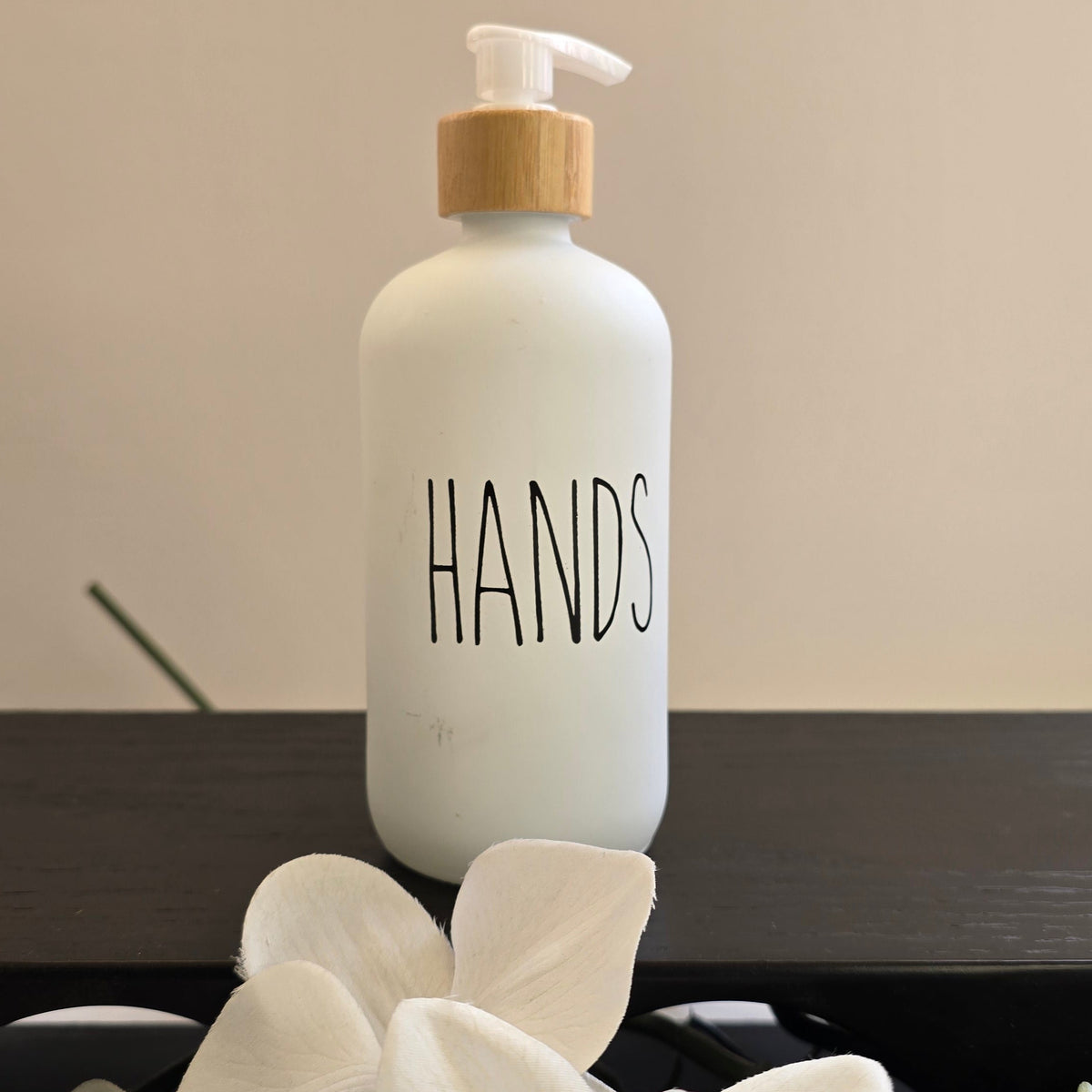 A frosted white hand sanitizer bottle with a bamboo pump dispenser and the word 'HANDS' printed on the front, styled on a dark surface with white orchid flowers in the foreground.