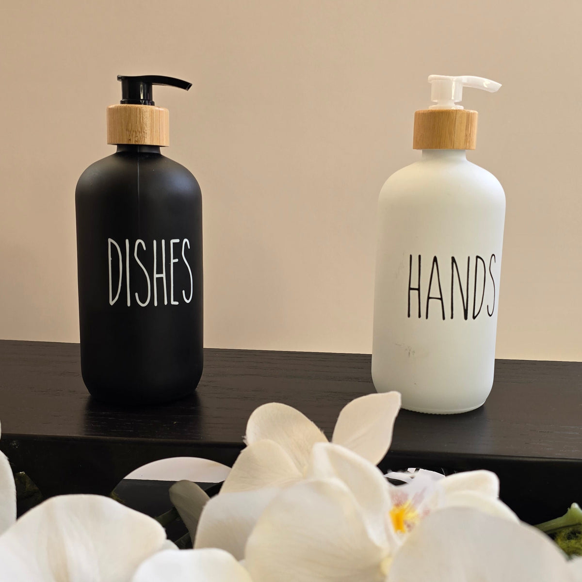 Two pump bottles labeled 'DISHES' and 'HANDS' sitting on a dark shelf, with white orchid flowers in the foreground against a beige wall.