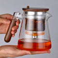 Glass teapot with wooden handle and lid held by a hand, filled with red liquid.