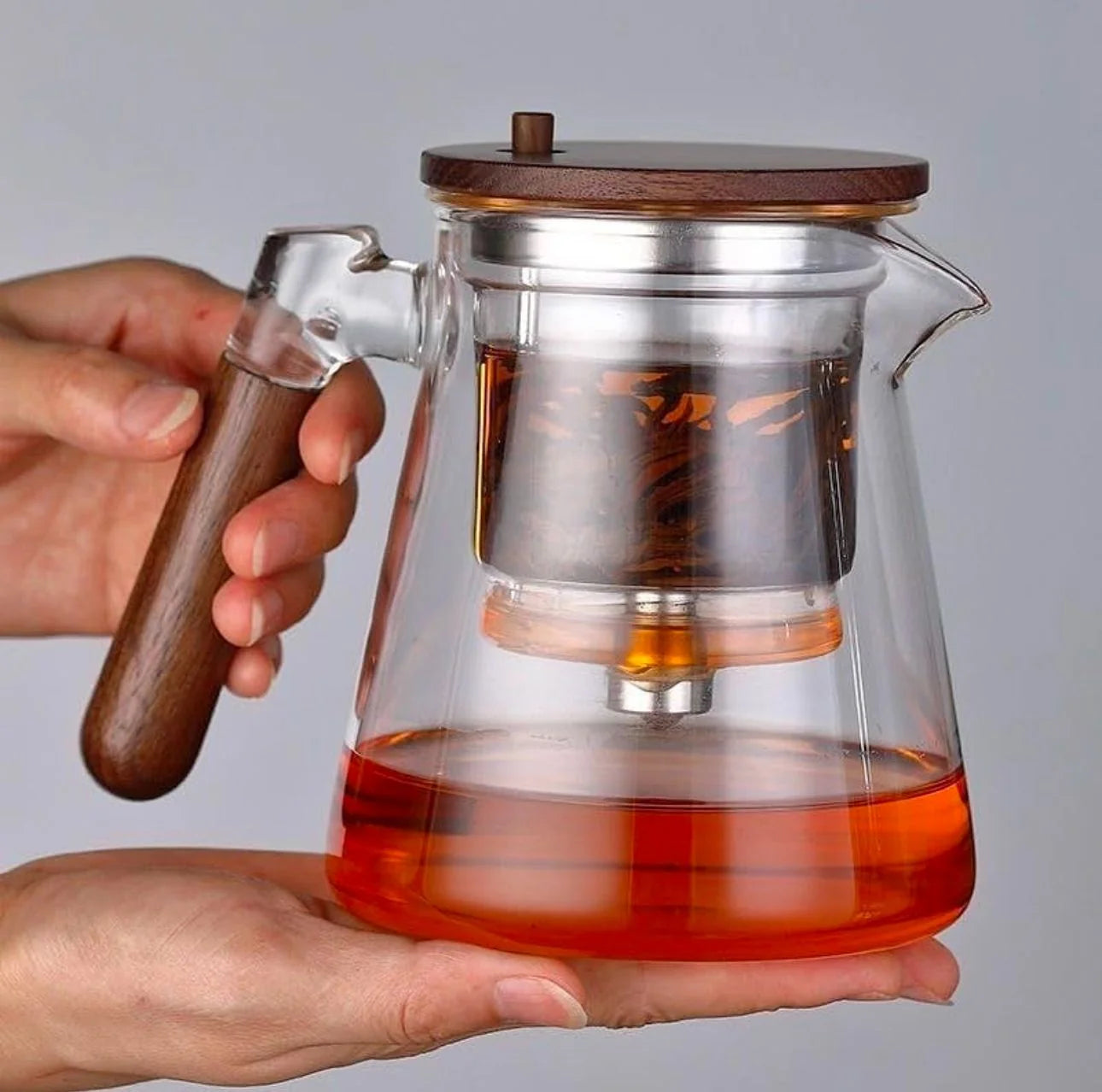 Glass teapot with wooden handle and lid held by a hand, filled with red liquid.
