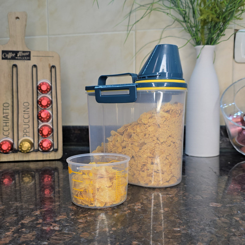 A clear plastic food storage container with a blue lid and handle, filled with brown granola or cereal, sits on a dark granite kitchen countertop next to a smaller clear container with yellow food. A Nespresso coffee capsule dispenser and a vase with flowers are visible in the background.