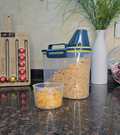 A clear plastic food storage container with a blue lid and handle, filled with brown granola or cereal, sits on a dark granite kitchen countertop next to a smaller clear container with yellow food. A Nespresso coffee capsule dispenser and a vase with flowers are visible in the background.