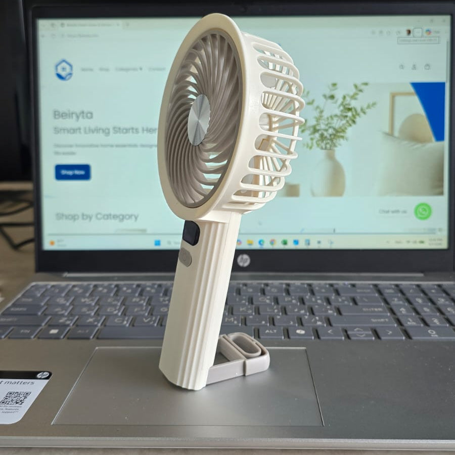 A small handheld fan in a cream color stands on a laptop palm rest, with a laptop screen visible in the background.