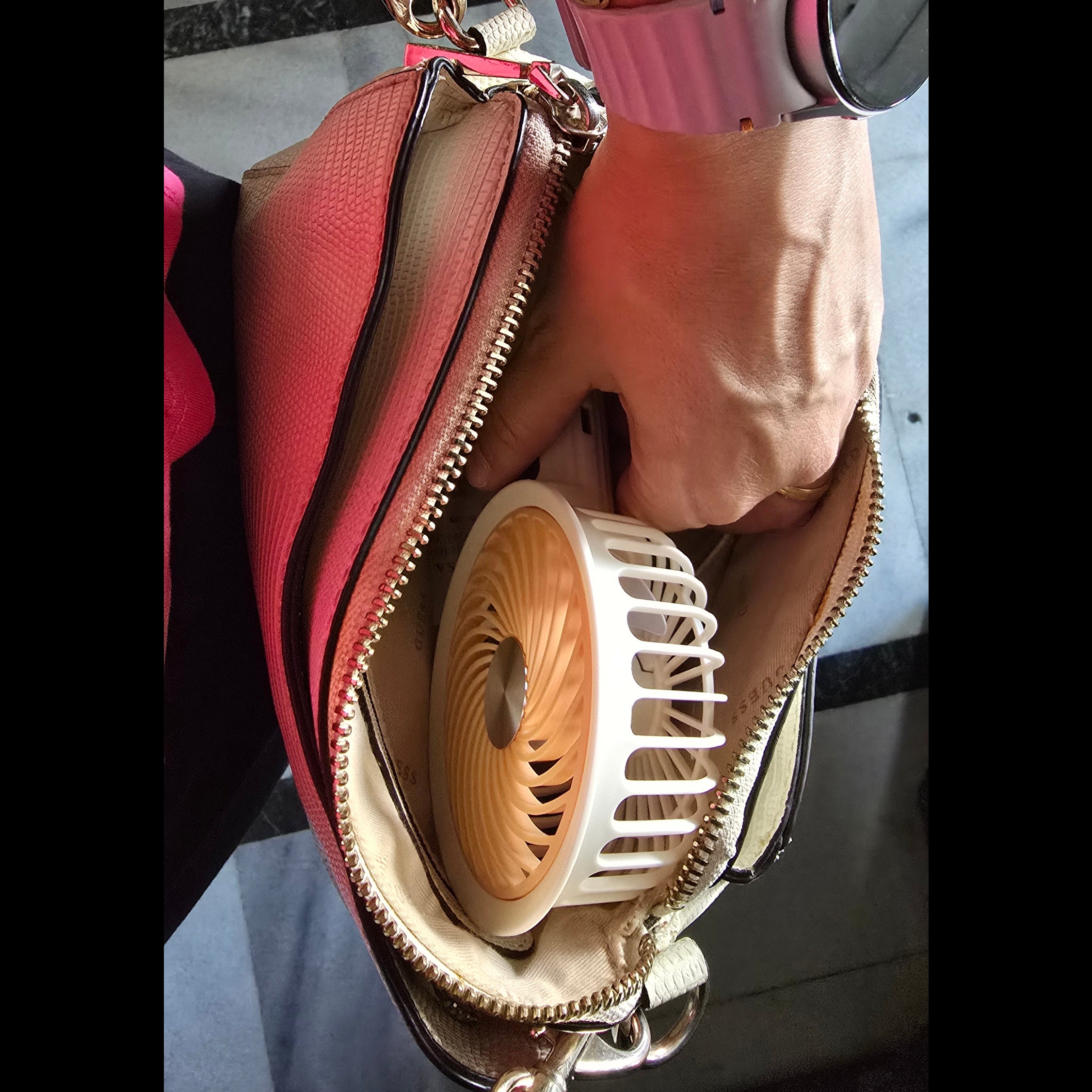 A close-up of a hand placing a small white and beige portable fan into a pink textured handbag with gold-tone zippers.