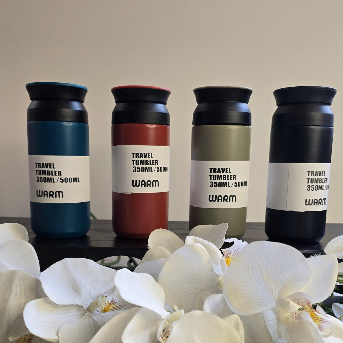 Four insulated travel tumblers with black lids and white labels reading 'TRAVEL TUMBLER 350ML 8oz WARM' are lined up on a dark surface, with a foreground of white orchids flowers.