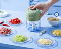 A hand is pressing the lid of a small electric food chopper, which has a sage green top and a clear blue container. The chopper is set on a light blue table surrounded by plates of chopped ingredients, including red chili peppers, minced meat, celery, garlic, and a bowl of soup, with its charging cable nearby.