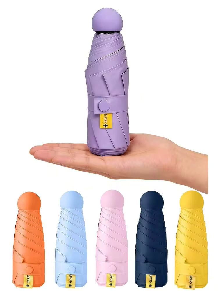 A compact, folded umbrella in light purple is shown resting on a person's hand to demonstrate its small size. Five additional folded umbrellas in orange, light blue, pink, navy, and yellow are lined up below on a white background.