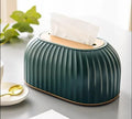 A ribbed dark teal tissue box cover with a light wood top and a white tissue protruding, styled on a gray tabletop with a plant and small dish in the background.