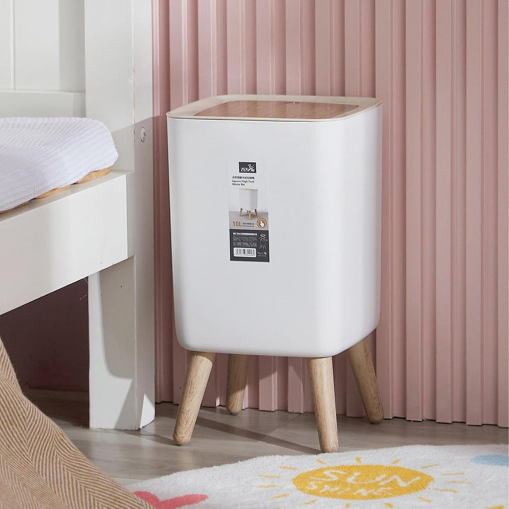 A modern white floor bin with a light wood-grain lid and four tapered wooden legs, styled in a bedroom next to a bed with a pink ribbed wall background.