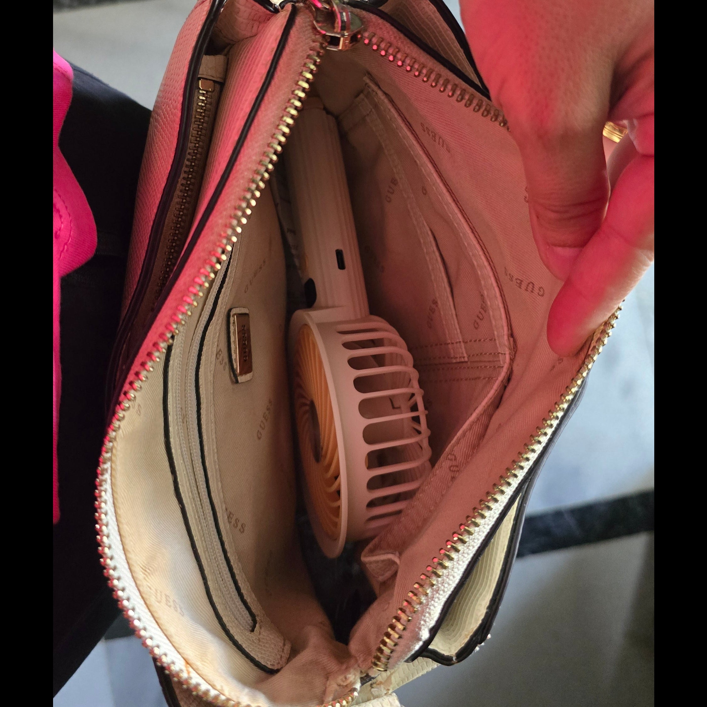 A close-up of a pink and beige handbag being held open by a person, showing the interior compartments and a small pink personal fan tucked inside. A pink wristwatch is also visible on the person's wrist.