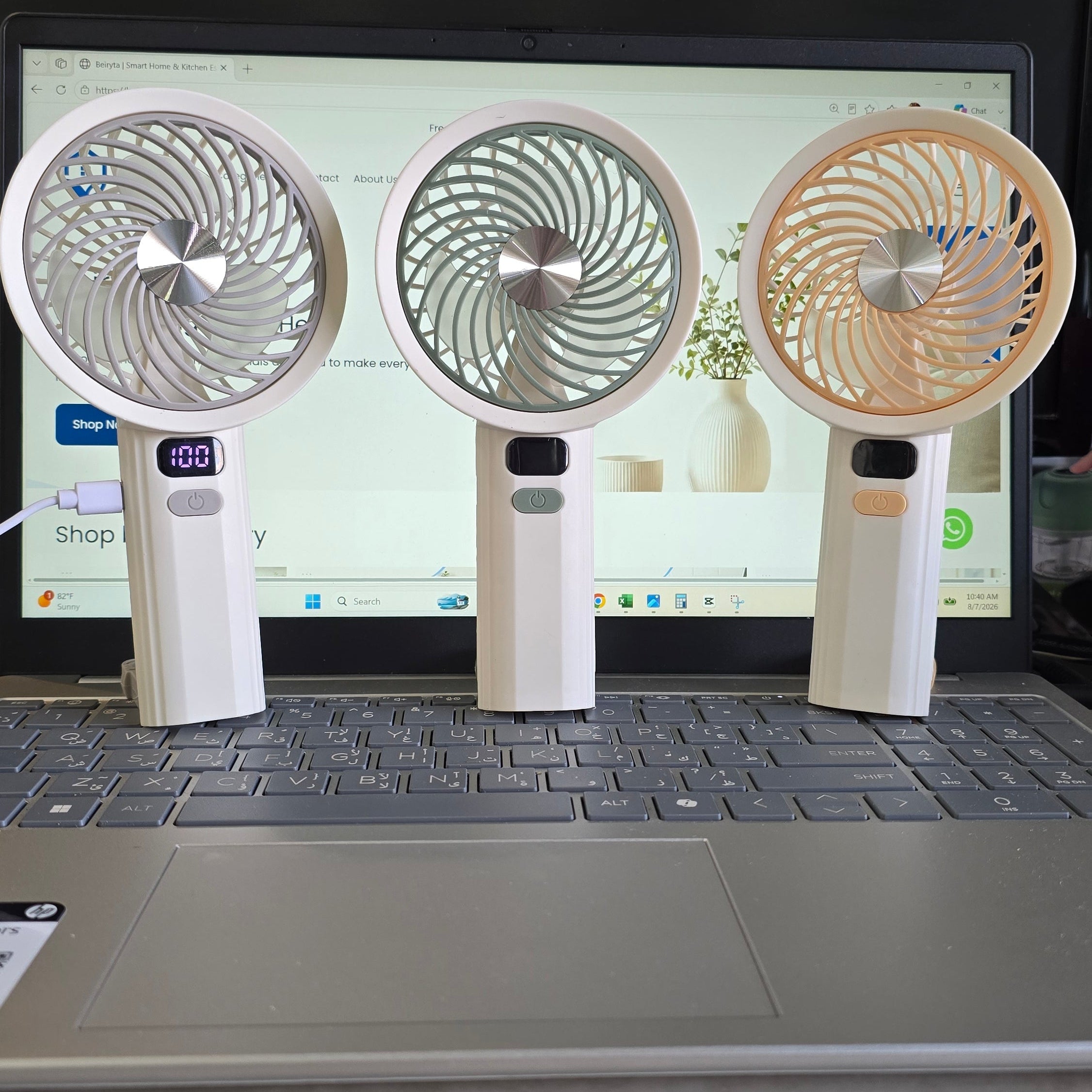 Three small handheld desk fans in white, silver, and beige standing on top of a laptop keyboard in an office setting.