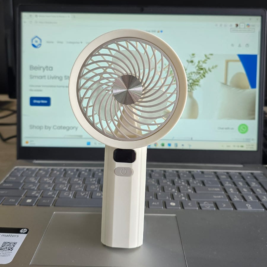 A small white handheld fan with a circular blade guard stands on a laptop keyboard. The laptop screen is on in the background, showing a webpage, and the fan is photographed in a real indoor setting on a desk.