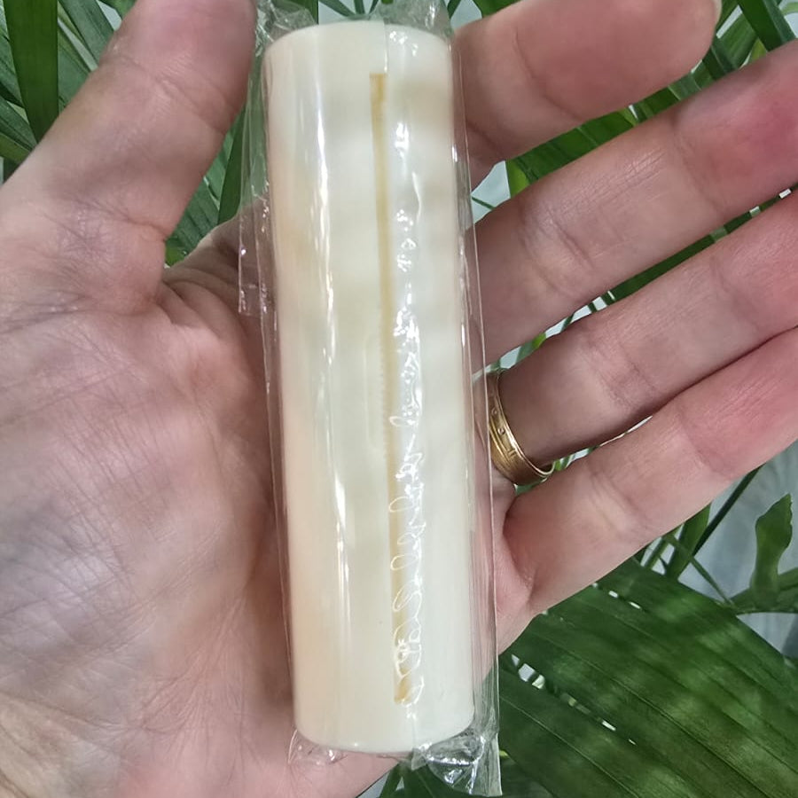 A hand holding a cylindrical, off-white soap bar wrapped in clear plastic, set against a background of green palm leaves.