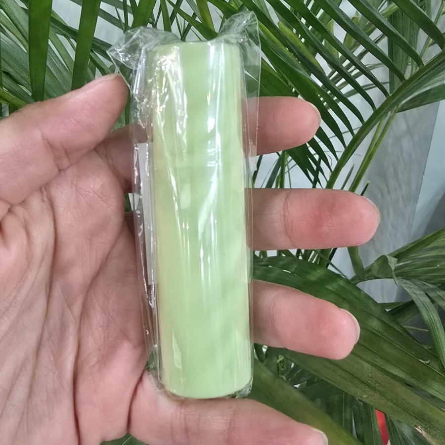 A hand holding a roll of pale green tape or bandage wrapped in clear plastic, with green plant leaves in the background.