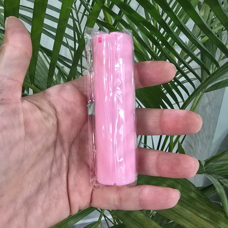 A hand holding a pink cylindrical product wrapped in clear plastic, positioned in front of a green potted plant.