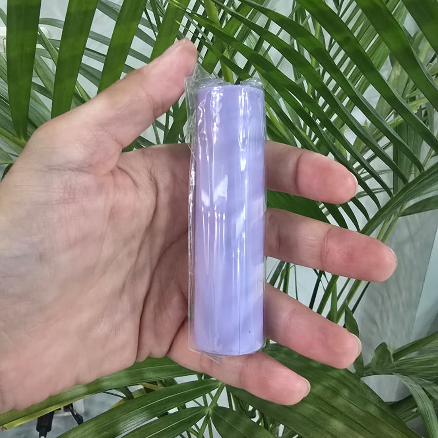 A hand holding a purple cylindrical candle wrapped in clear plastic, set against a background of green palm leaves.