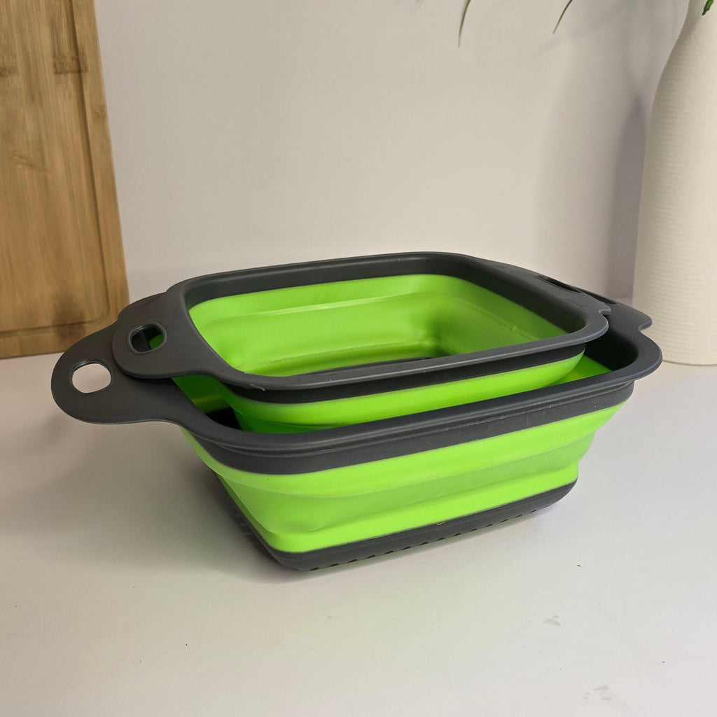 Two collapsible silicone bowls, one lime green and one dark gray, stacked on a white tabletop with a white wall and a potted plant in the background.