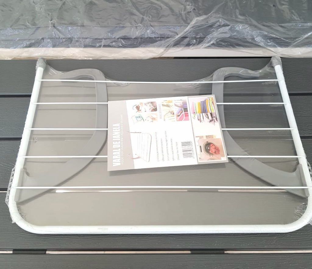 A white metal clothesline with gray plastic feet, shown wrapped in protective plastic with its instruction booklet and product card on top, set against a dark gray slatted background.