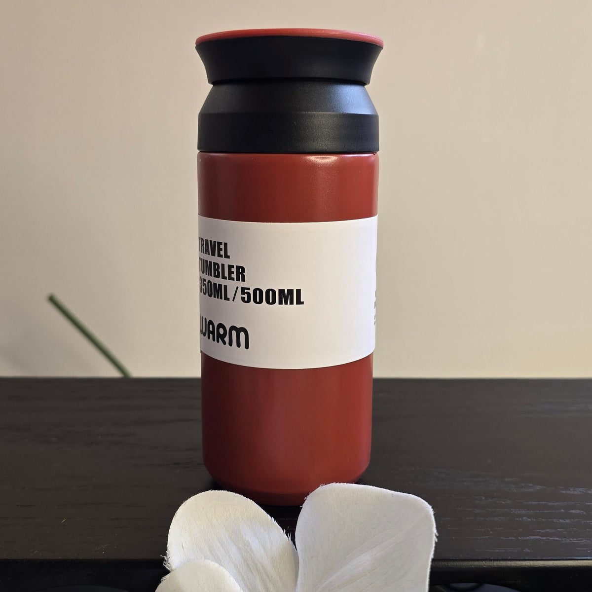 A 500ml travel tumbler with a terracotta red body, black lid, and a white label, displayed on a dark surface with white flowers in the foreground.