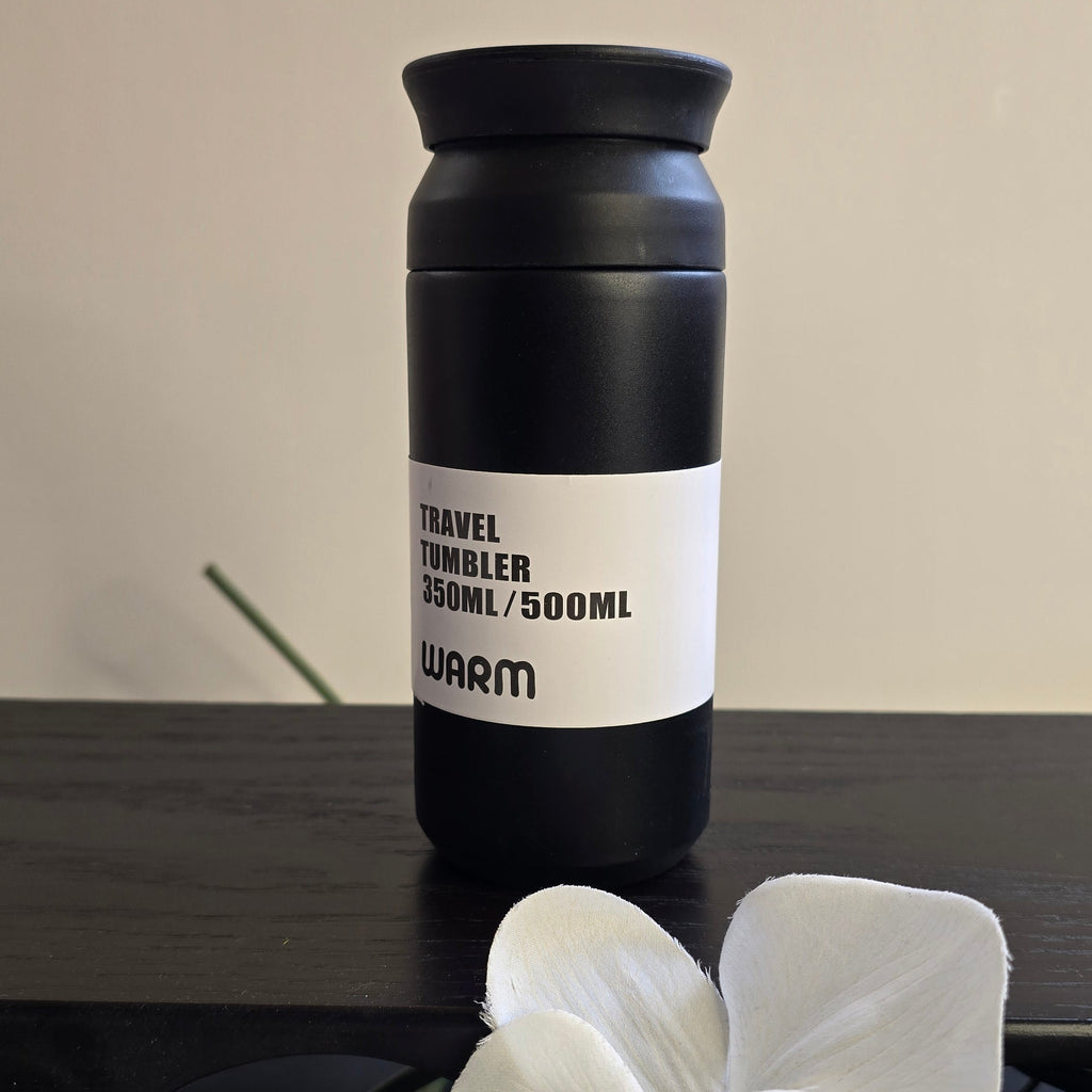 A matte black travel tumbler with a white label reading 'TRAVEL TUMBLER 350ML/500ML' and 'WARM', placed on a dark wooden surface with white orchid flowers in the foreground and a beige wall behind.