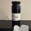 A matte black travel tumbler with a white label reading 'TRAVEL TUMBLER 350ML/500ML' and 'WARM', placed on a dark wooden surface with white orchid flowers in the foreground and a beige wall behind.