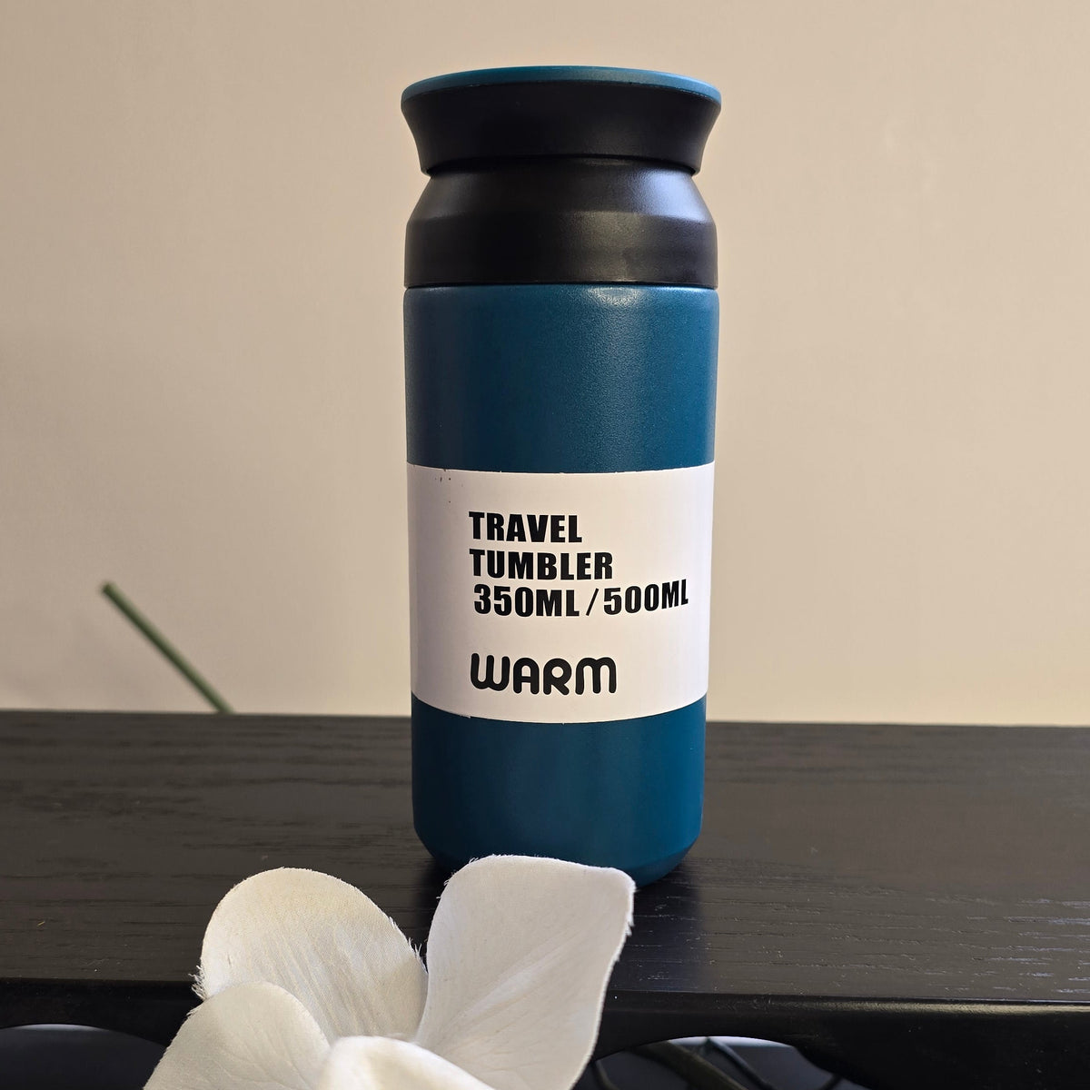 A 350ml travel tumbler with a dark blue body, black lid, and a white label that reads 'TRAVEL TUMBLER 350ML/500ML WARM'. The tumbler is placed on a dark wooden surface with white orchids in the foreground and a beige wall in the background.