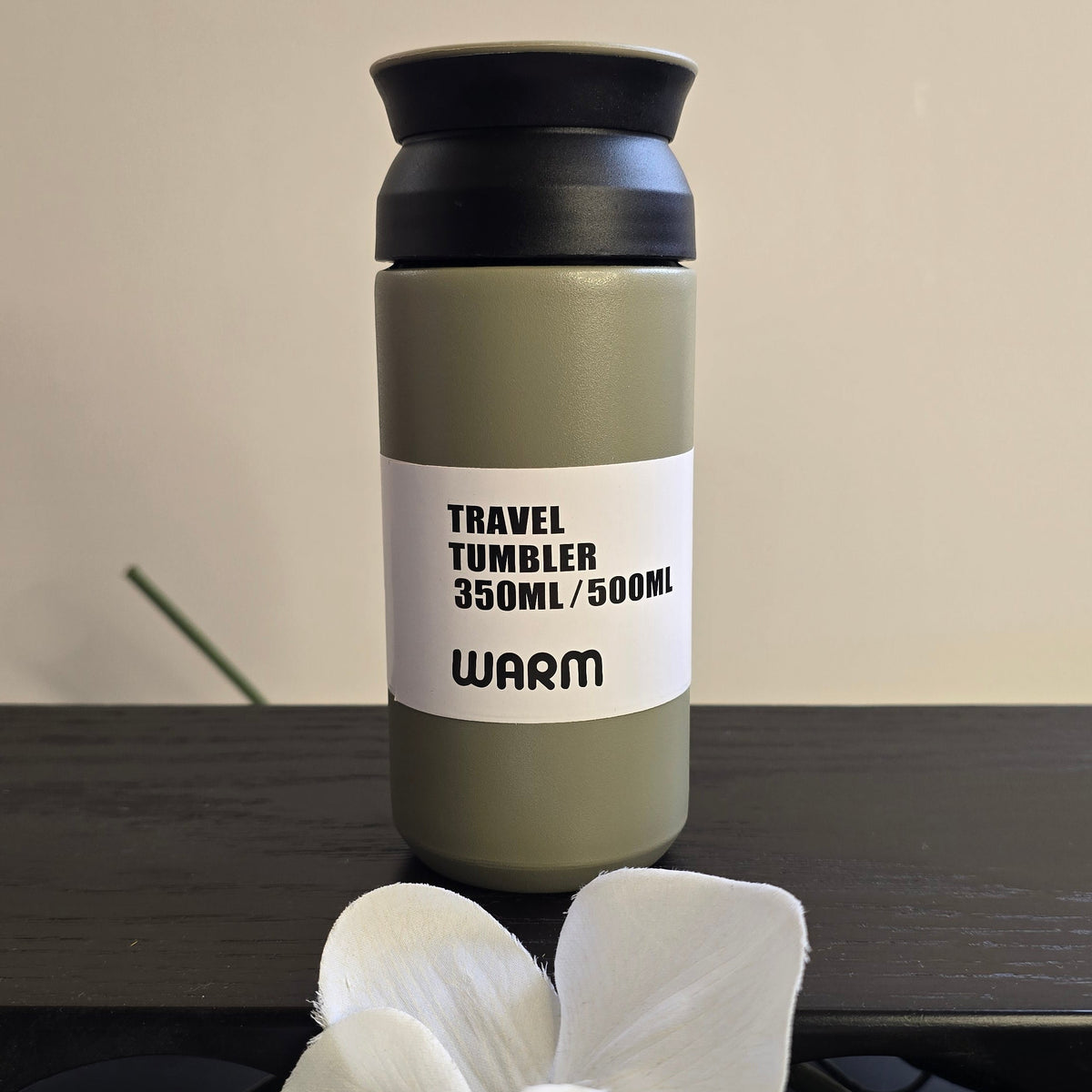 A 350ml travel tumbler with an olive green body, black lid, and a white label that reads 'TRAVEL TUMBLER 350ML/500ML WARM'. The tumbler is placed on a dark wooden surface with white orchid flowers in the foreground and a beige wall in the background.
