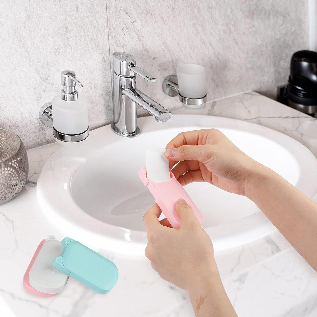 A person's hands are shown using a pink and white toilet paper holder over a white ceramic bathroom sink. Another matching holder in light blue and white lies on the marble countertop nearby.