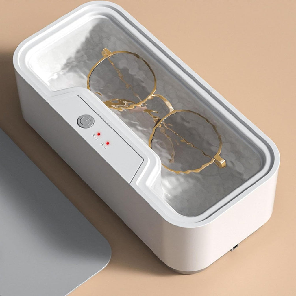 A white rectangular glass UV lamp or jewelry cleaner with a transparent lid, shown with a pair of gold-rimmed glasses inside. The device has a control panel with a power button and red indicator lights on the front, set against a warm beige background.