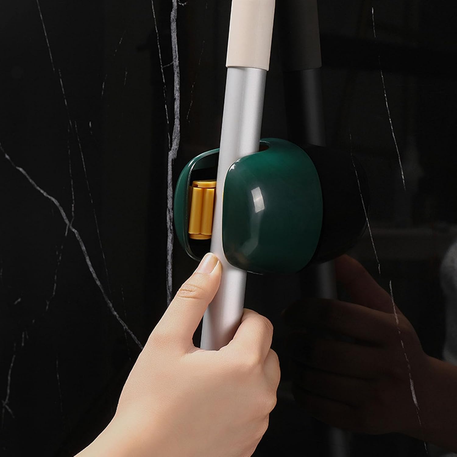 A close-up of a hand holding a silver pole against a black marble wall, with a dark green and black wall-mounted hook holding the pole in place.