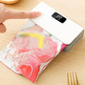 A hand is pressing down on the white lid of a vacuum sealer device, which is sealing a clear plastic bag containing pink and yellow food, on a light wood tabletop.